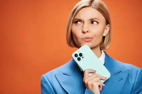 Спантеличена молода жінка з коротким волоссям позує зі своїм смартфоном на помаранчевому фоні — стокове фото