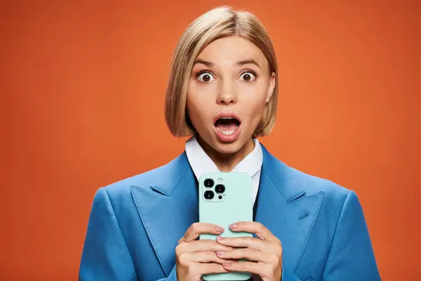 Surprised young woman with blonde short hair posing with her smartphone on orange background — Stock Photo