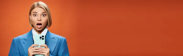 Shocked young woman with blonde short hair posing with her smartphone on orange background, banner — Stock Photo