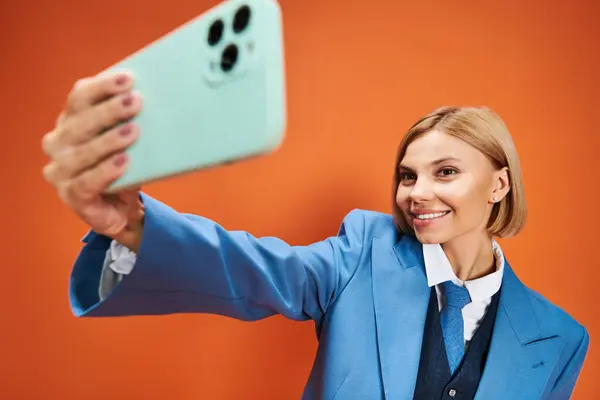 Glückliche gut gekleidete Frau mit kurzen blonden Haaren in schicker Kleidung mit Telefon auf orangefarbenem Hintergrund — Stockfoto