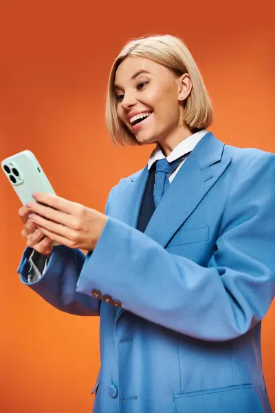 Joyeuse femme sophistiquée avec des cheveux blonds courts en tenue chic tenant le téléphone sur fond orange — Photo de stock