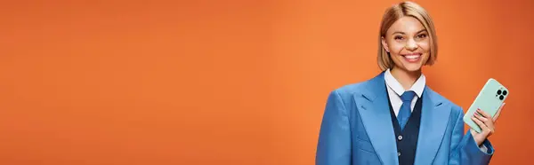 Mujer elegante feliz con pelo corto y rubio en traje elegante sosteniendo el teléfono sobre fondo naranja, pancarta - foto de stock