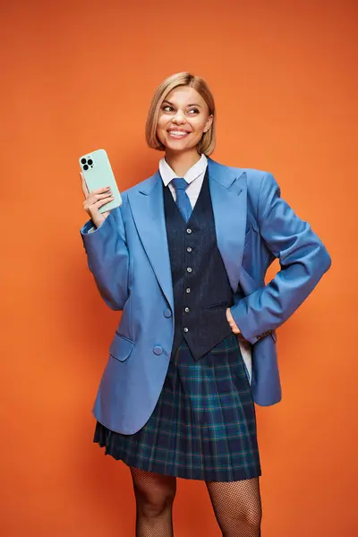 Joyful sophisticated woman with short blonde hair in chic attire holding phone on orange backdrop — Stock Photo