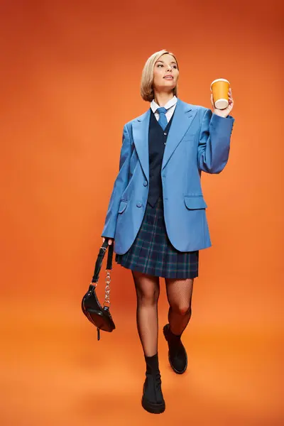 Mujer sofisticada alegre con pelo corto rubio sosteniendo bolso y café sobre fondo naranja - foto de stock