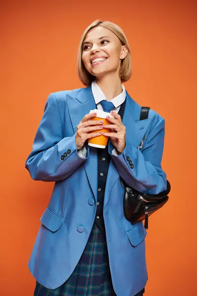 Alegre mujer sofisticada con pelo corto rubia sosteniendo bolso y café sobre fondo naranja - foto de stock