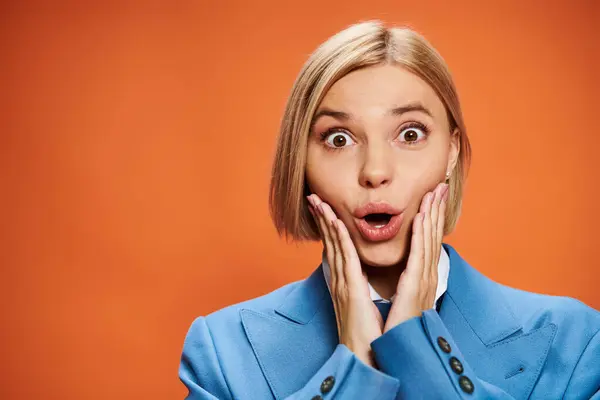 Shocked young woman with short blonde hair in sophisticated attire posing on orange backdrop — Stock Photo