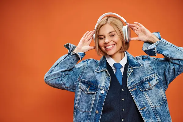 Mujer alegre y elegante con pelo rubio con auriculares en chaqueta de mezclilla posando sobre fondo naranja - foto de stock