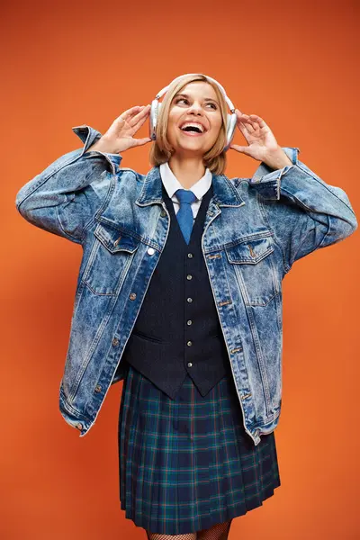 Joyful stylish woman with blonde hair with headphones in denim jacket posing on orange backdrop — Stock Photo