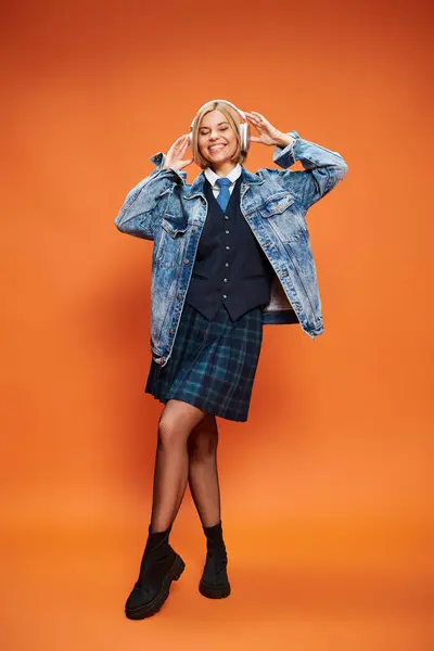 Positive appealing woman with blonde hair with headphones in denim jacket posing on orange backdrop — Stock Photo