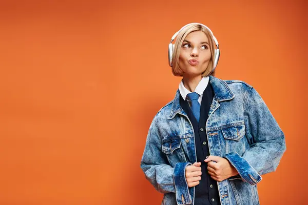 Cheerful appealing woman with blonde hair with headphones in denim jacket posing on orange backdrop — Stock Photo