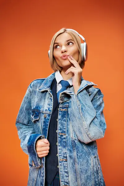 Щаслива молода жінка з світлим волоссям з навушниками в джинсовій куртці позує на помаранчевому тлі — стокове фото