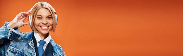 Merry young woman with blonde hair with headphones in denim jacket posing on orange backdrop, banner — Stock Photo