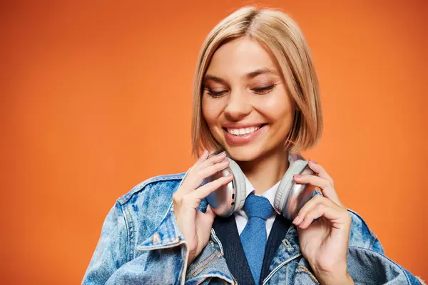 Jeune femme positive avec des cheveux blonds avec des écouteurs dans la veste en denim posant sur fond orange — Photo de stock