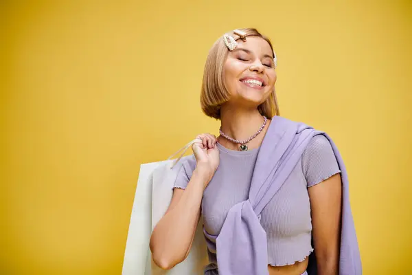 Contented woman with accessories in chic attire posing with shopping bag on yellow backdrop — Stock Photo