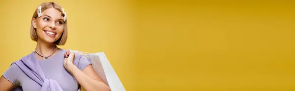 Happy woman with accessories in chic attire posing with shopping bag on yellow backdrop, banner — Stock Photo
