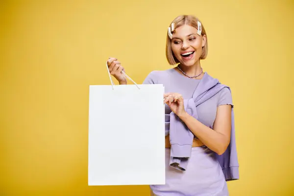 Joyful attractive woman with accessories in chic attire posing with shopping bag on yellow backdrop — Stock Photo
