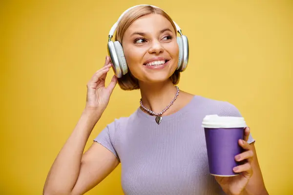 Cheerful stylish woman with headphones enjoying music and hot coffee while posing on yellow backdrop — Stock Photo