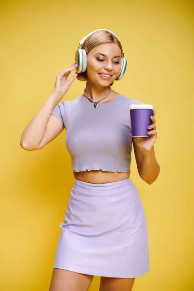 Mujer alegre y elegante con auriculares disfrutando de la música y el café caliente mientras posa en el fondo amarillo - foto de stock