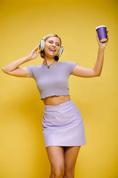 Mujer de moda contenta con auriculares disfrutando de la música y el café caliente mientras posa en el fondo amarillo - foto de stock