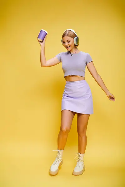 Glad trendy woman with headphones enjoying music and hot coffee while posing on yellow backdrop — Stock Photo