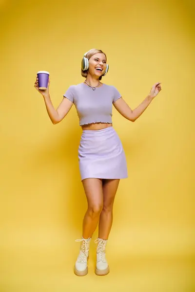 Alegre mujer de moda con auriculares disfrutando de la música y el café caliente mientras posa en el fondo amarillo - foto de stock