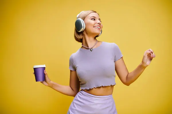 Alegre mujer de moda con auriculares disfrutando de la música y el café caliente mientras posa en el fondo amarillo - foto de stock