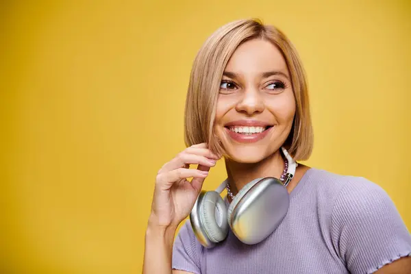 Encantadora mujer alegre con pelo corto y rubio y auriculares disfrutando de la música en el fondo amarillo - foto de stock