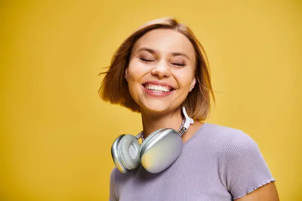 Hübsche fröhliche Frau mit kurzen blonden Haaren und Kopfhörern, die Musik auf gelbem Hintergrund genießt — Stockfoto