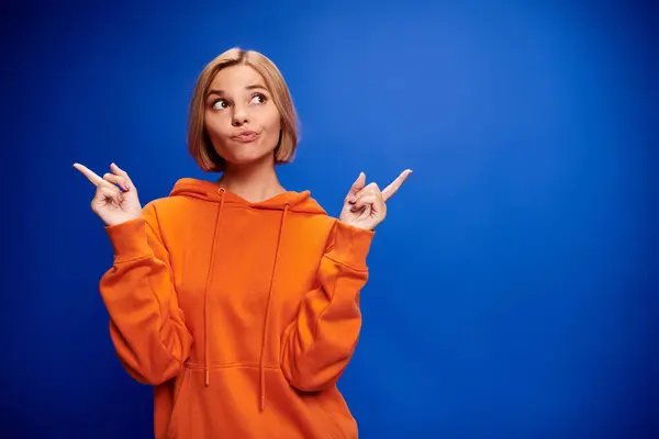 Glad blonde woman with short hair in vibrant orange hoodie posing actively on blue backdrop — Stock Photo