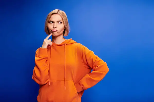 Alluring puzzled blonde woman with short hair in stylish orange hoodie posing on blue background — Stock Photo