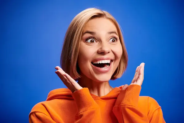 Upbeat blonde woman with short hair in vibrant orange hoodie posing actively on blue backdrop — Stock Photo