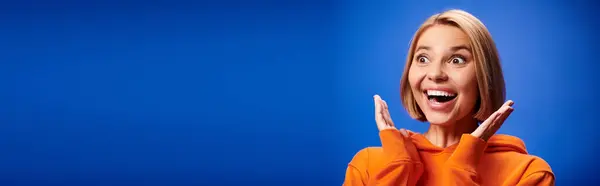 Positive woman with short hair in vibrant orange hoodie posing actively on blue backdrop, banner — Stock Photo