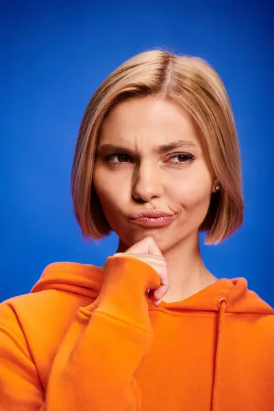Attractive puzzled blonde woman with short hair in stylish orange hoodie posing on blue background — Stock Photo