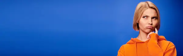 Puzzled blonde woman with short hair in trendy orange hoodie posing on blue background, banner — Stock Photo