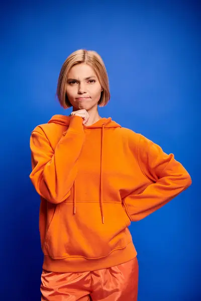 Confusa mujer rubia atractiva con pelo corto en sudadera con capucha naranja elegante posando sobre fondo azul - foto de stock