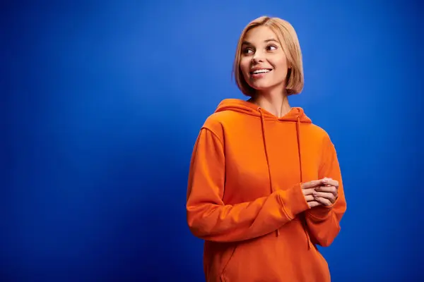 Attraktive, fröhliche Frau mit kurzen Haaren in leuchtend orangefarbenem Kapuzenpulli posiert aktiv vor blauem Hintergrund — Stock Photo