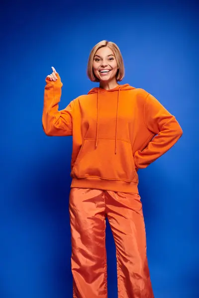 Attractive cheerful woman with short hair in vibrant orange hoodie posing actively on blue backdrop — Stock Photo