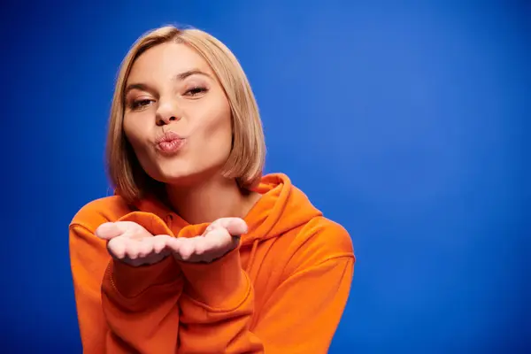 Mujer alegre bien vestida con pelo corto en sudadera con capucha naranja vibrante posando activamente sobre fondo azul - foto de stock