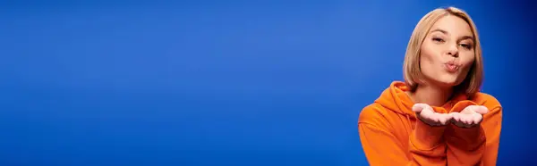 Cheerful woman with short hair in vibrant orange hoodie posing actively on blue backdrop, banner — Stock Photo