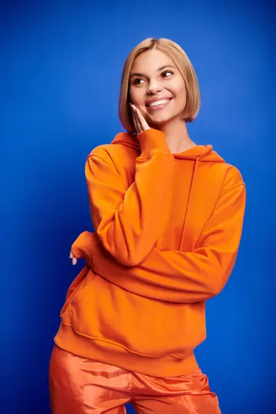 Polished cheerful woman with short hair in vibrant orange hoodie posing actively on blue backdrop — Stock Photo