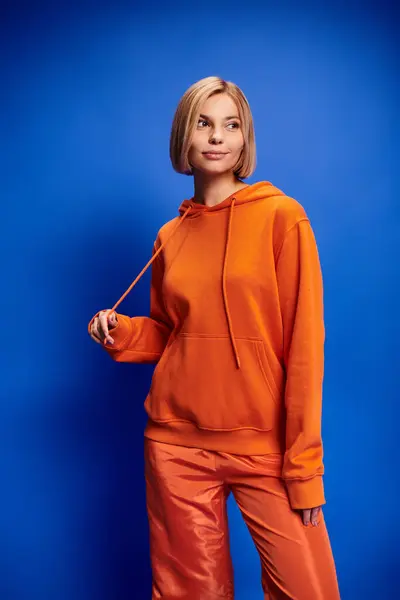 Exquisite cheerful woman with short hair in vibrant orange hoodie posing actively on blue backdrop — Stock Photo