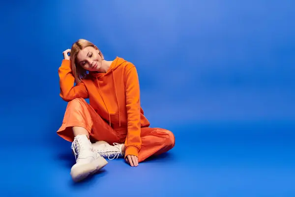 Mujer alegre de buen gusto con el pelo corto en sudadera con capucha naranja vibrante posando activamente sobre fondo azul — Stock Photo