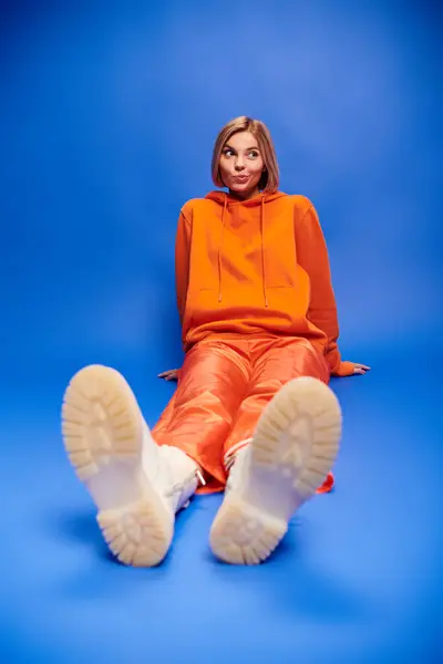 Graziosa donna allegra con i capelli corti in vibrante felpa arancione con cappuccio in posa attiva su sfondo blu — Stock Photo