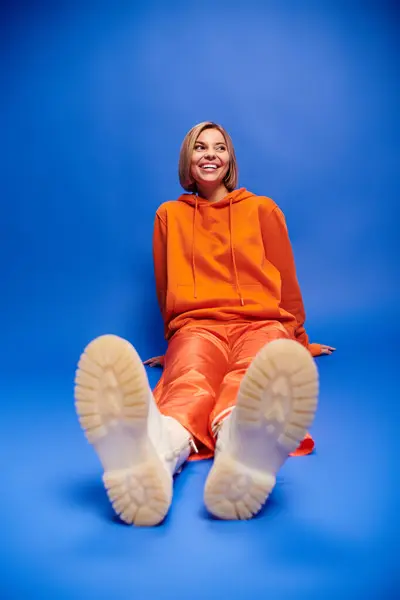 Trendige, fröhliche Frau mit kurzen Haaren in leuchtend orangefarbenem Kapuzenpulli posiert aktiv vor blauem Hintergrund — Stockfoto