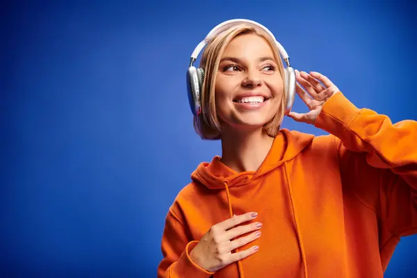 Attraktive, fröhliche Frau mit kurzen blonden Haaren und Kopfhörern, die Musik auf blauem Hintergrund genießt — Stockfoto