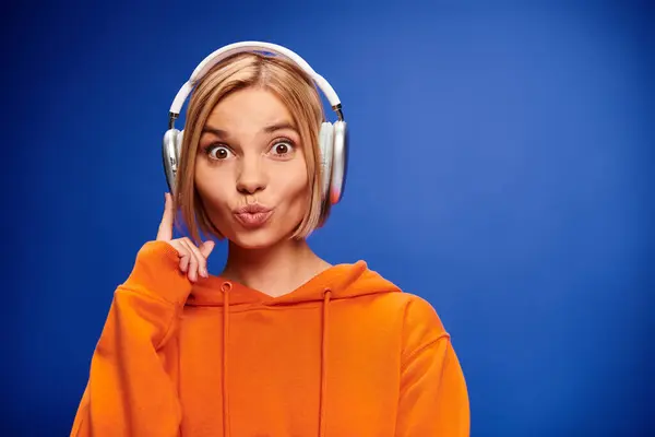 Attraktive stilvolle Frau mit kurzen blonden Haaren und Kopfhörern, die Musik auf blauem Hintergrund genießt — Stockfoto