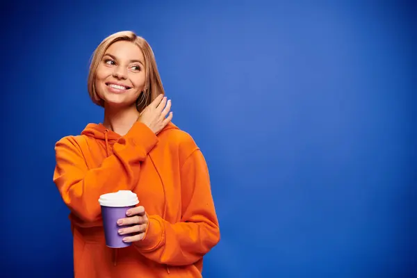Atractiva mujer alegre con pelo corto y rubio en ropa vibrante sosteniendo taza de café sobre fondo azul - foto de stock