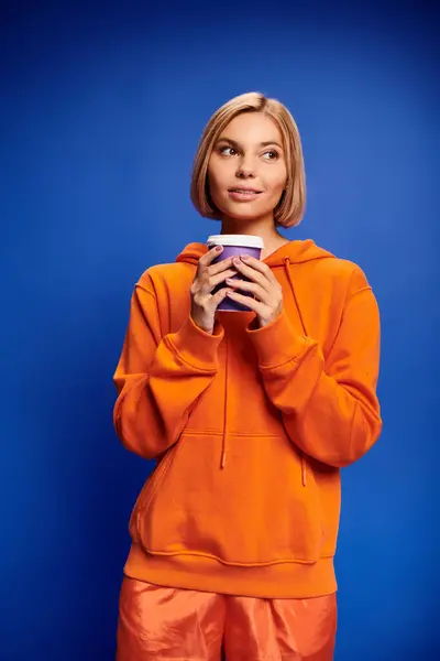 Atractiva mujer alegre con pelo corto y rubio en ropa vibrante sosteniendo taza de café sobre fondo azul - foto de stock