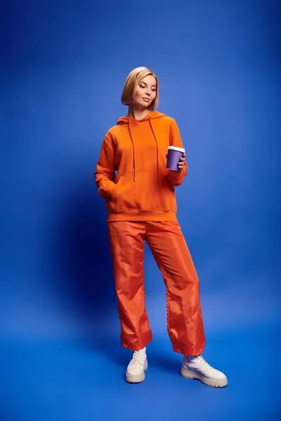 Alluring joyous woman with short blonde hair in vibrant clothes holding coffee cup on blue backdrop — Stock Photo