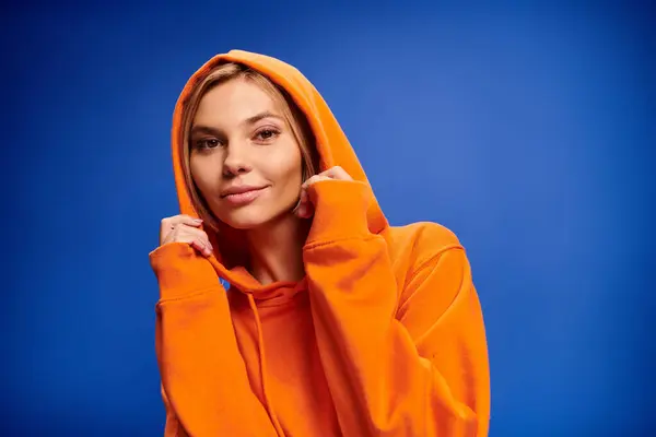 Beautiful jolly woman with blonde short hair in vivid attire with hood on posing on blue backdrop — Stock Photo